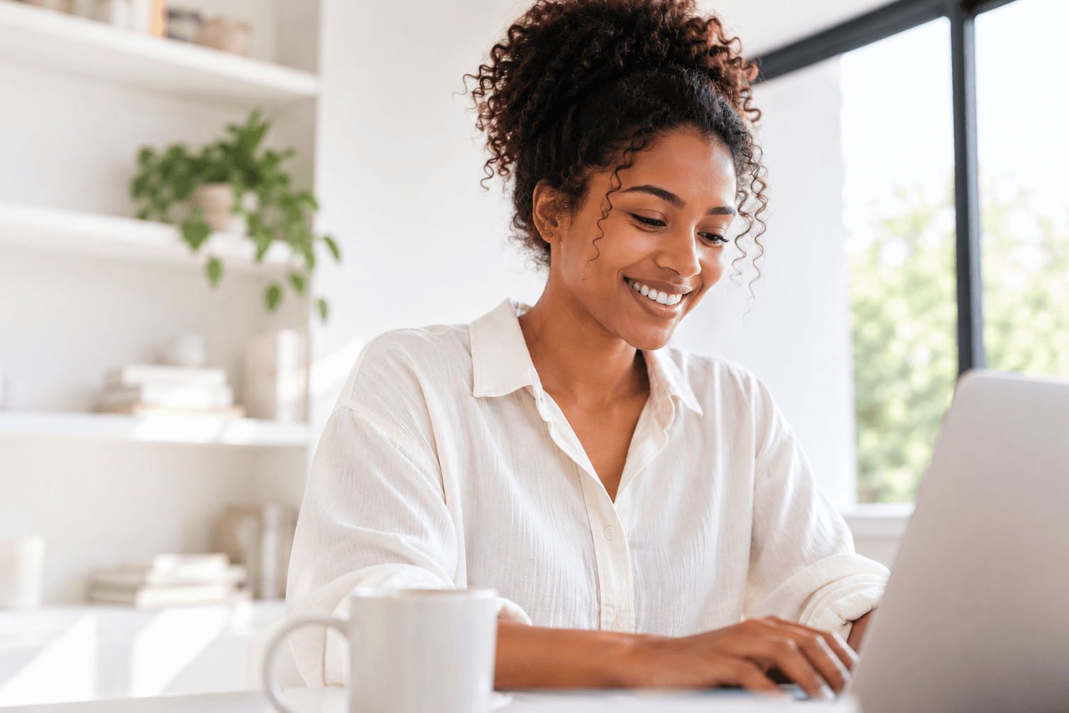 A woman smiling while working on a laptop in a bright room
